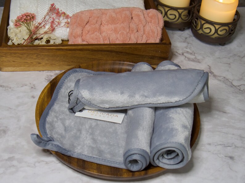 May include: Three gray microfiber makeup remover cloths rolled up and placed on a wooden plate. The cloths are labeled "Delicada Beauty".