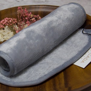 May include: A gray makeup remover cloth rolled up on a wooden surface. The cloth has a gray border and is labeled "Delicada Beauty".