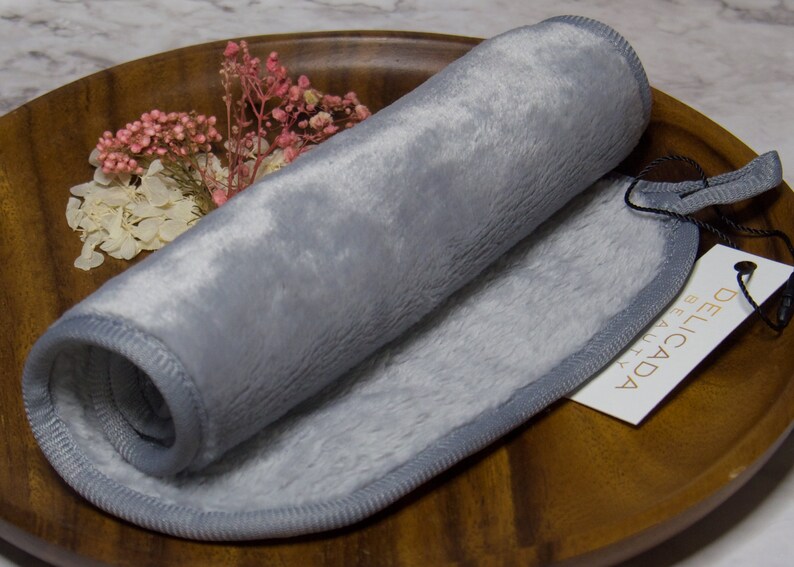 May include: A gray makeup remover cloth rolled up on a wooden surface. The cloth has a gray border and is labeled "Delicada Beauty".