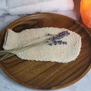 May include: A beige, fish-shaped bath mitt with a textured surface, resting on a wooden plate. A sprig of lavender is placed on top. A white towel and a glowing orange salt lamp are in the background.