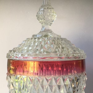 May include: A clear glass candy dish with a diamond pattern and a red band around the top. The lid has a clear glass finial.