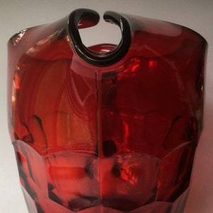 May include: A red glass pitcher with a unique, curved handle and a faceted design. The pitcher has a wide base and a rounded body.