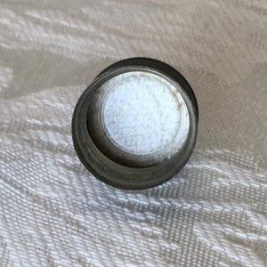 May include: A small, cylindrical metal object with a white, fluffy interior. The object has a dark, metallic exterior and a circular opening. The item is likely a component of a larger product.