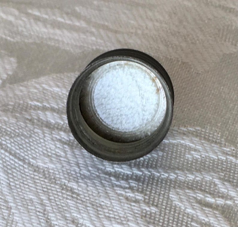 May include: A small, cylindrical metal object with a white, fluffy interior. The object has a dark, metallic exterior and a circular opening. The item is likely a component of a larger product.