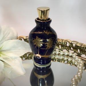 Op de afbeelding: Een donkerblauwe parfumfles met een gouden dop en accenten. De fles heeft gouden palmboomontwerpen en de woorden "Hawaiian Night Mist". De fles wordt gepresenteerd op een reflecterend oppervlak.