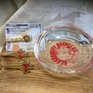 May include: A clear glass ashtray with a red and white graphic that says "Electricity Recycles". Also, a prop $100 bill, a gold-colored tie clip with a cowboy hat, and two gold-colored tie clips with a red figure.