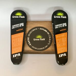 May include: Two black and orange Green Flash IPA tap handles with the text "Tangerine Soul Style" and "Citra Cascade". A wooden coaster with the Green Flash logo and website is centered between the handles.