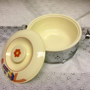 May include: A round, cream-colored casserole dish with a matching lid. The lid features a central orange flower design and a colorful fruit basket graphic. The dish is set within a polished chrome metal frame with cut-out details and side handles.
