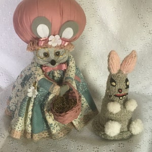 May include: A gray mouse wearing a pink hat and a blue dress holds a pink basket. A gray knitted rabbit with white accents sits next to the mouse.