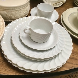 May include: White ceramic dinnerware set with a scalloped edge design. The set includes plates, saucers, and teacups. The teacups have a spiral design. The dinnerware is arranged on a wooden surface.