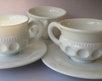 May include: Three white milk glass cups and saucers with a raised dot pattern. The cups have a rounded shape and a single handle.