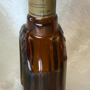 May include: A small, brown glass bottle with a metal cap. The bottle has a label that reads "1.5 fl oz." The bottle's shape is rectangular with rounded edges. The bottle is likely for a liquid product.