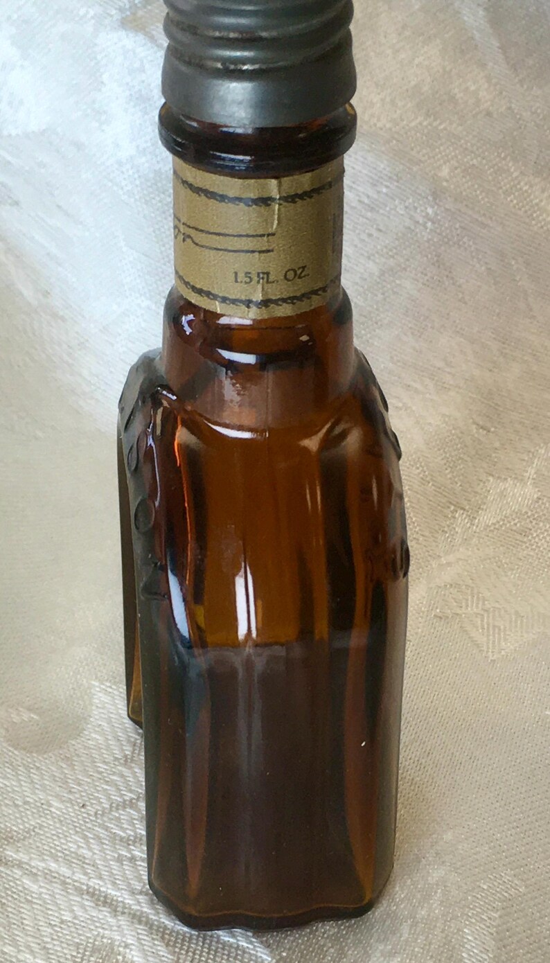 May include: A small, brown glass bottle with a metal cap. The bottle has a label that reads "1.5 fl oz." The bottle's shape is rectangular with rounded edges. The bottle is likely for a liquid product.