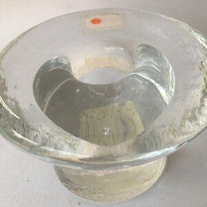 May include: A clear glass candle holder with a wide, flat base and a raised, circular center. The holder has a slightly textured surface and a small sticker on the side.