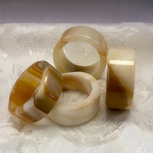 May include: A collection of four smooth, polished stone rings featuring translucent bands in shades of cream, beige, and warm amber. Each ring displays unique natural banding and color variations within the polished mineral material.