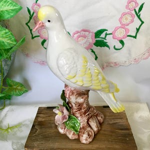 May include: A white ceramic dove figurine with yellow and pink accents. The bird is perched on a brown tree trunk base with pink flowers and green leaves. The dove has a yellow crest and pink beak.