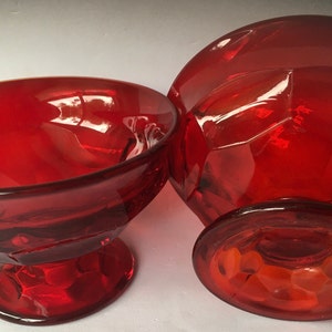 May include: Two red glass bowls with a faceted design. The bowls have a wide, round base and a smaller, round top.