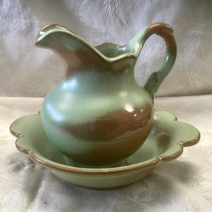 May include: A ceramic pitcher and matching basin set featuring a smooth, glossy glaze in blended shades of sage green and warm brown. The pitcher has a curved handle and a scalloped rim, while the basin features a matching scalloped edge design.