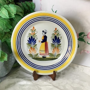 May include: A decorative ceramic plate with a yellow and blue rim. The plate features a central design of a woman in traditional attire, flanked by stylized floral motifs in blue, orange, and green. The plate is displayed on a wooden stand.