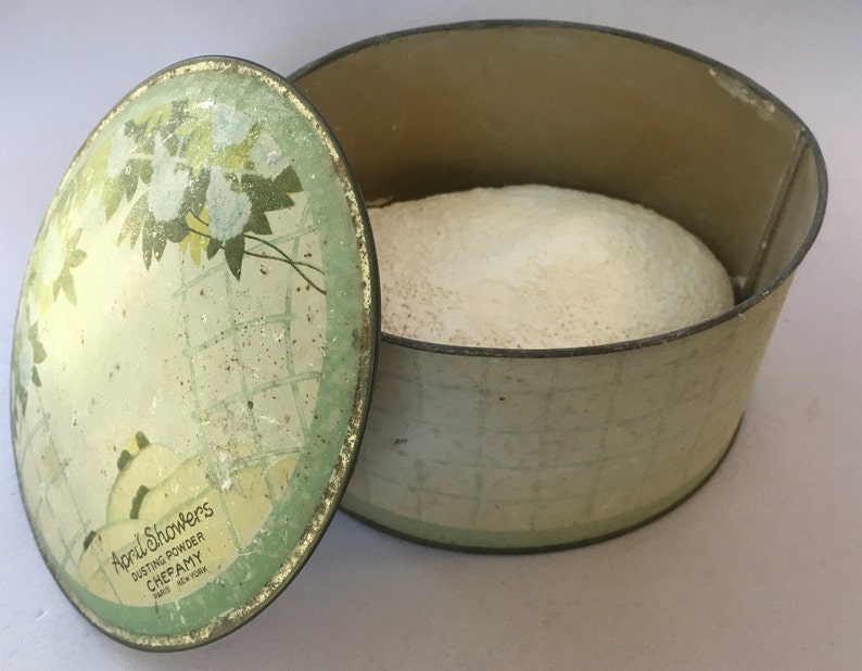 May include: Vintage green and white tin box with a floral design on the lid. The box contains a white powder and the lid reads "April Showers Dusting Powder Chepamy New York".