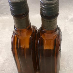 May include: Two amber-colored glass bottles with dark metal caps. The bottles have a textured design and are labeled with text. Each bottle appears to hold 0.5 fl oz of liquid. The bottles are likely vintage or antique.