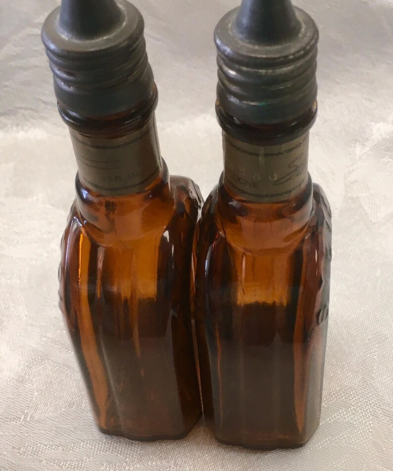 May include: Two amber-colored glass bottles with dark metal caps. The bottles have a textured design and are labeled with text. Each bottle appears to hold 0.5 fl oz of liquid. The bottles are likely vintage or antique.