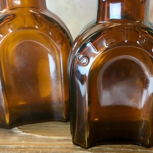 May include: Two antique amber glass bottles with a rounded rectangular shape. The word "STETS" is embossed on the top of each bottle. The bottles have a vintage aesthetic and are likely for storing liquids.