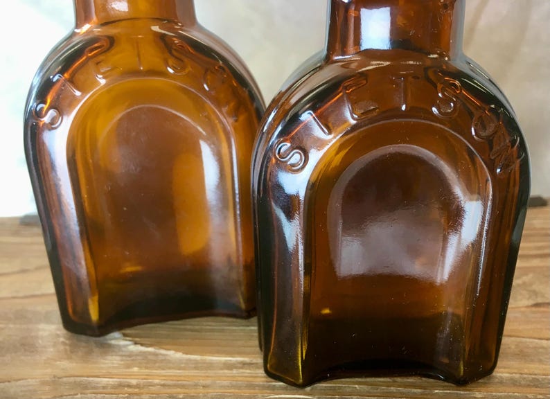May include: Two antique amber glass bottles with a rounded rectangular shape. The word "STETS" is embossed on the top of each bottle. The bottles have a vintage aesthetic and are likely for storing liquids.