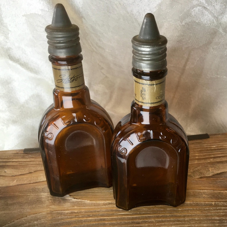 May include: Two vintage brown glass bottles with metal stoppers. The bottles have a rounded shape with the word "Stetson" embossed on the front. Each bottle has a label near the top. The bottles are displayed on a wooden surface.