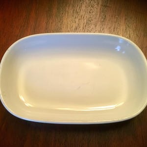 May include: A white, oval-shaped serving dish with rounded corners. The dish has a smooth, glossy surface and is set against a dark wooden background. The dish is empty and appears to be made of ceramic or porcelain.