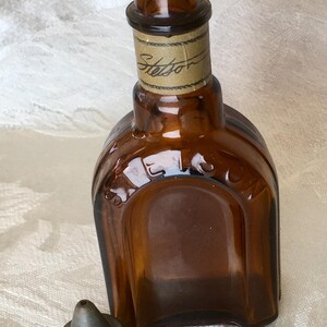 May include: A vintage brown glass bottle with a metal cap. The bottle has a rectangular shape with rounded edges and the word "Stetson" is printed on a label around the neck. The cap is a dull silver color. The bottle is empty.