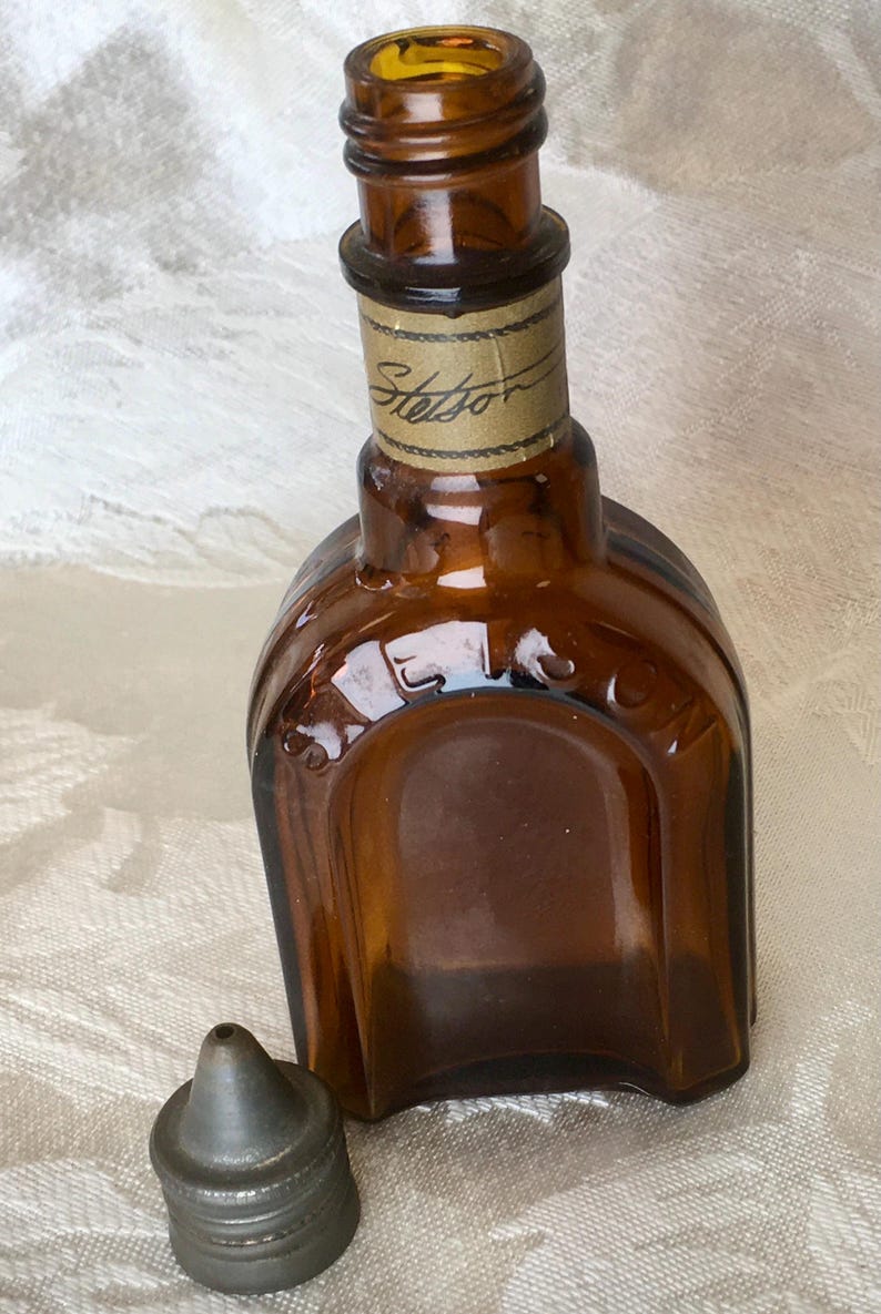May include: A vintage brown glass bottle with a metal cap. The bottle has a rectangular shape with rounded edges and the word "Stetson" is printed on a label around the neck. The cap is a dull silver color. The bottle is empty.