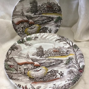 May include: Two ceramic plates with a rural landscape design. The plates feature a black and white illustration of a village scene with thatched-roof houses, a horse-drawn cart, and figures. The plates have a brown and white color scheme.