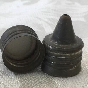 May include: Two antique metal bottle stoppers. One is a cylindrical cap with ridges, and the other is a conical stopper with a small hole at the top. Both are dark gray with signs of wear, suggesting age and use.