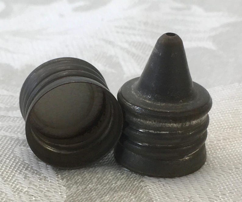 May include: Two antique metal bottle stoppers. One is a cylindrical cap with ridges, and the other is a conical stopper with a small hole at the top. Both are dark gray with signs of wear, suggesting age and use.