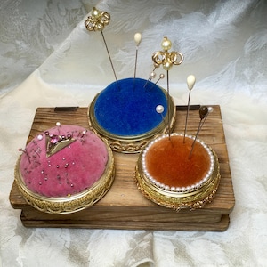 May include: Three antique pin cushions in gold-toned metal frames, each with a velvet top in pink, blue, and orange. The cushions are adorned with decorative pins, some with pearl and jewel accents, and sit on a wooden base.