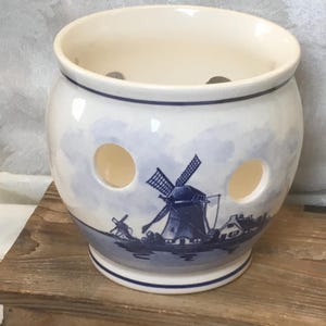 May include: Cream-colored ceramic garlic keeper with a blue and white windmill design. The round keeper has three circular ventilation holes and a blue rim. The windmill scene is painted on the front of the keeper.