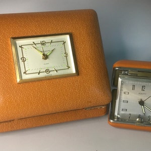 May include: Two vintage orange leather travel alarm clocks. The larger clock has a white face with black hands and the text "Alarm" printed on the face. The smaller clock has a white face with black hands and the text "Equity" printed on the face.
