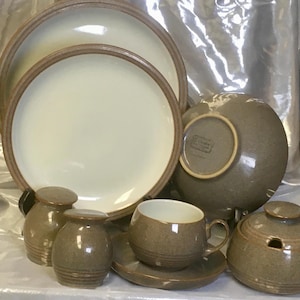 May include: A stoneware dinnerware set featuring two large plates with cream centers and brown rims, a bowl, a teacup with a saucer, a lidded sugar bowl, and two shakers. The pieces have a speckled brown finish and a smooth, glazed appearance.