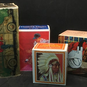 May include: Four vintage aftershave boxes. The boxes are all different colors and have different designs. One box features a car, another a woman in a red dress, another a Native American chief, and the last one a white ceramic pipe.