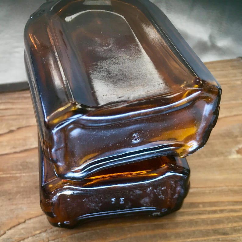 May include: Two amber-colored glass bottles stacked on top of each other. The bottles have a rectangular shape with rounded edges and a slightly curved top. The glass has a glossy finish, and the bottles appear to be vintage.