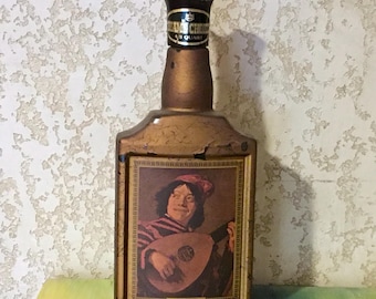 May include: A vintage glass bottle with a gold-colored label featuring a painting of a man playing a lute. The bottle has a black plastic cap with the text "Grand Choice 1/5 Quart".
