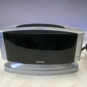 May include: A silver and black Sharp digital alarm clock. The clock has a large black display, with silver accents and a silver base. The word "SNOOZE" is printed on the top, and "SHARP" is printed on the front.