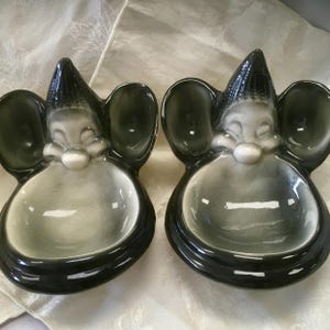 May include: Two ceramic gnome-shaped dishes with a black and gray color scheme. Each dish features a smiling gnome face with a pointed hat, large ears, and a bowl-shaped body. The dishes are likely decorative or for serving small items.