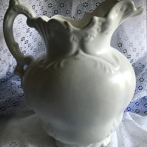 May include: White ceramic pitcher with a decorative floral design. The pitcher has a wide mouth and a handle.