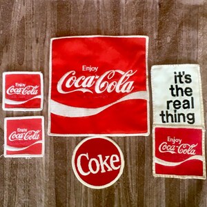 May include: A collection of six fabric patches featuring Coca-Cola branding. Includes four square red patches with white Enjoy Coca-Cola text, one circular red patch with white Coke text, and one rectangular white patch with black text it's the real thing.