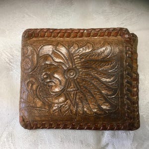 May include: A brown leather wallet with a detailed carved design featuring a Native American figure with a feathered headdress. The wallet has a braided border and a rectangular shape.