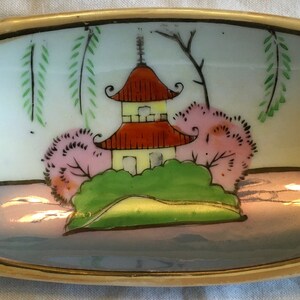 May include: A small, oval-shaped dish with a gold rim and a blue and white painted scene of a pagoda, trees, and a river. The dish is made of ceramic and has a glossy finish.