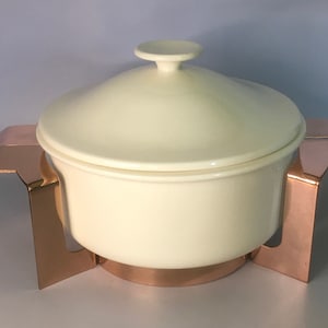 May include: A white ceramic serving dish with a lid, sitting on a copper stand. The stand has four legs and a round base.