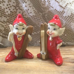 May include: Two ceramic elf figurines wearing bright red outfits and pointed hats. Each elf has a gold-colored heart accent on the chest and is holding a pair of brown wooden skis. The figures have white skin, pointed ears, and painted facial features.